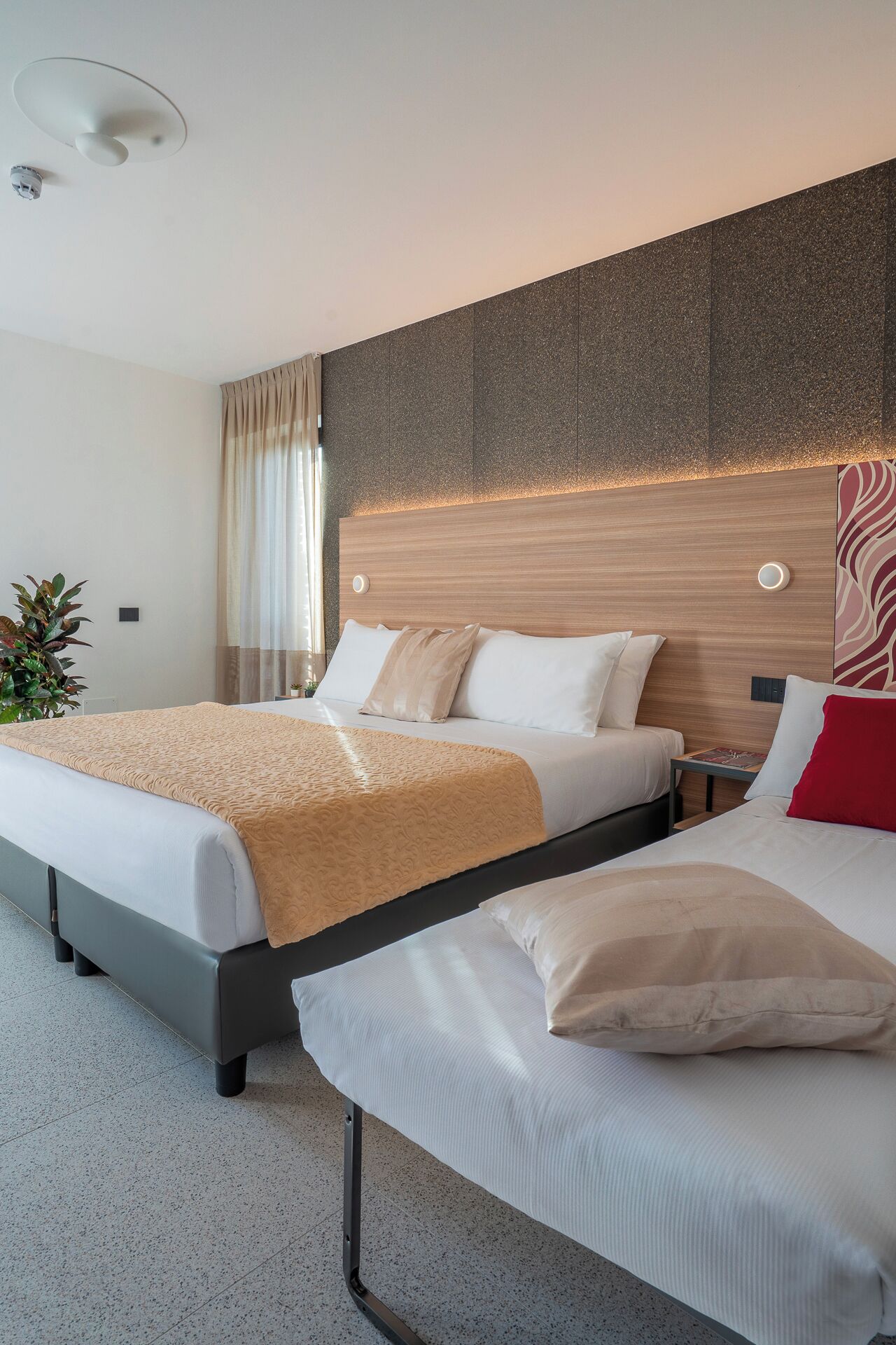 Ein Hotelzimmer mit einem großen Bett, einem kleineren Bett, einem Nachttisch, einer Pflanze und einem modernen Wanddesign.