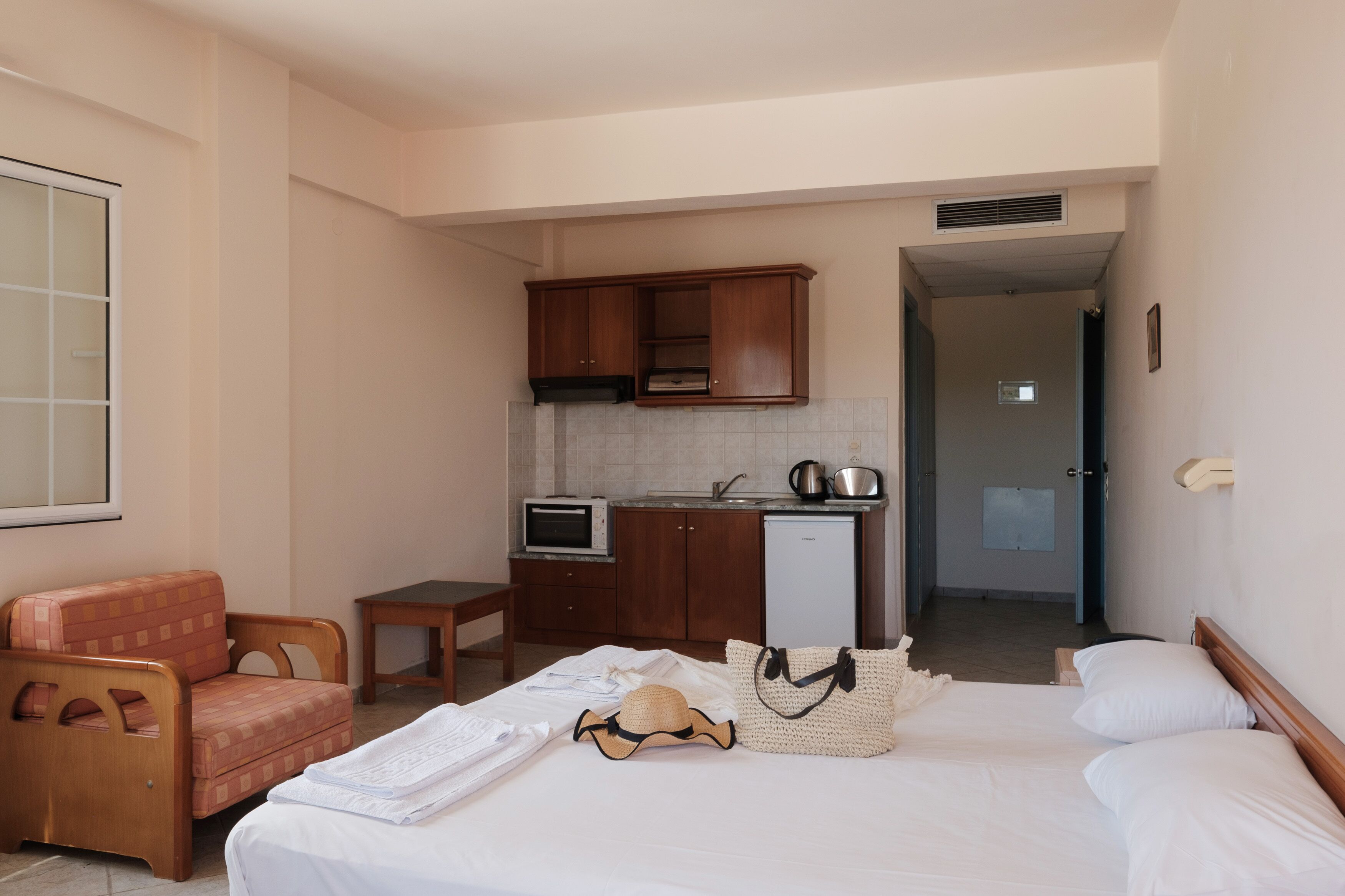 A compact hotel room with a bed, a small kitchenette, a wooden chair, and a view of the hallway.