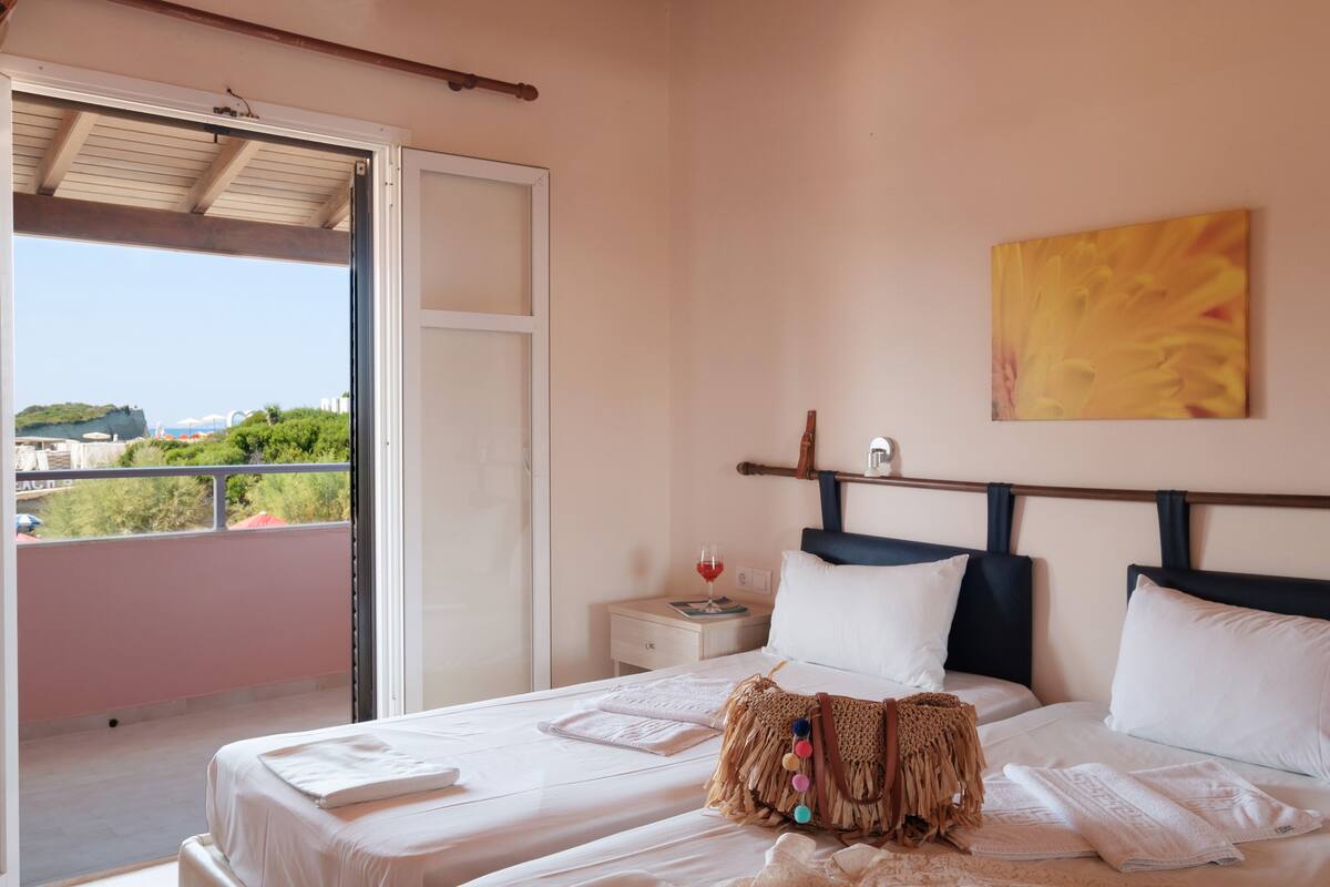 A bedroom with a bed, a nightstand, a window with a view of greenery and buildings, and a painting on the wall.
