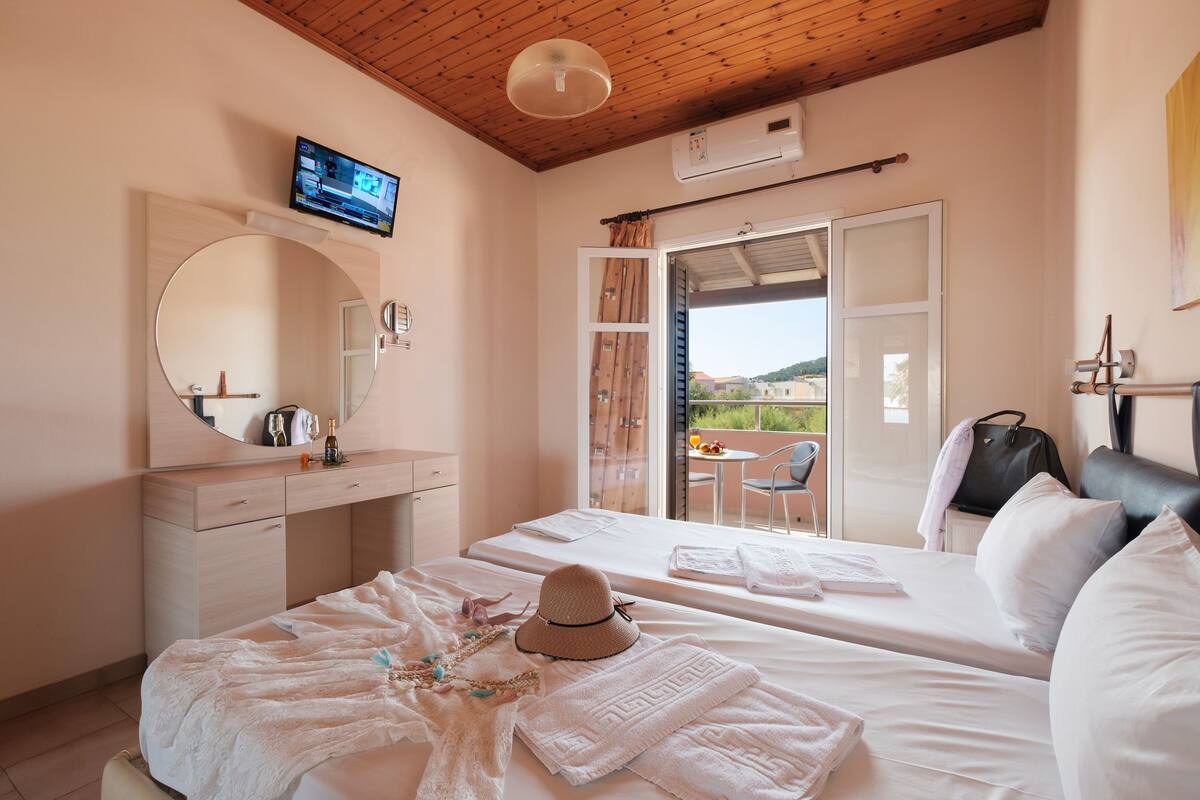 A hotel room with two beds, a TV, a mirror, a balcony with a table and chairs, and a view of the outside.