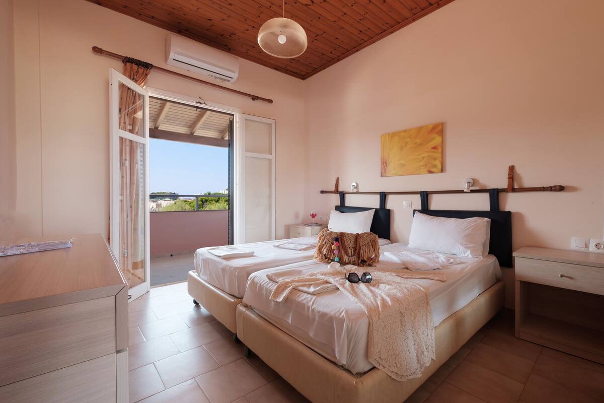 A bedroom with two beds, a wooden ceiling, an air conditioning unit, and a view of greenery outside.