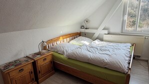 Quarto com teto inclinado, cama de madeira, mesa de cabeceira, cômoda e janela com vista para árvores.