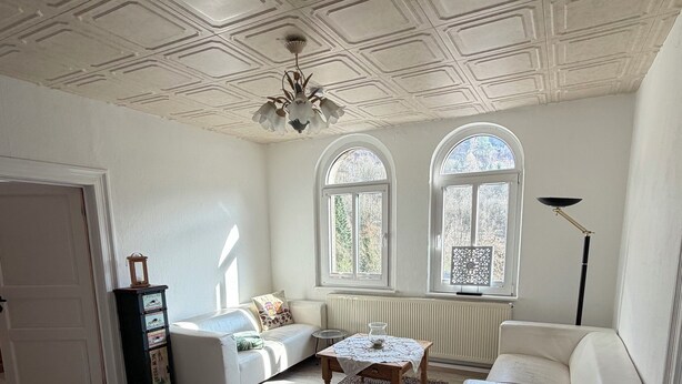 Uma sala de estar ampla com teto alto, duas janelas em arco, um sofá branco, uma mesa de centro e um abajur.