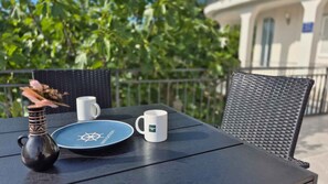 Ein Tisch mit einem Teller, einer Tasse und einer Vase auf einem Balkon mit Geländer und Grünpflanzen im Hintergrund.