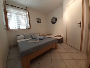 Une petite chambre propre, avec un lit simple, une table de chevet, une horloge et une fenêtre avec des stores.