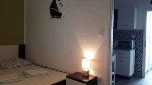 Ein Schlafzimmer mit einem Bett, einem Nachttisch und einem Beistelltisch mit einer Lampe. Im Hintergrund ist eine Küchenzeile mit einer Mikrowelle und Schränken zu sehen.