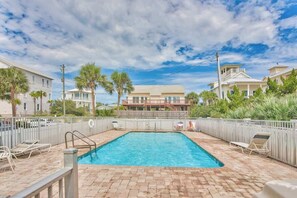 A rectangular swimming pool with a brick deck, surrounded by a white fence, and palm trees in the background.