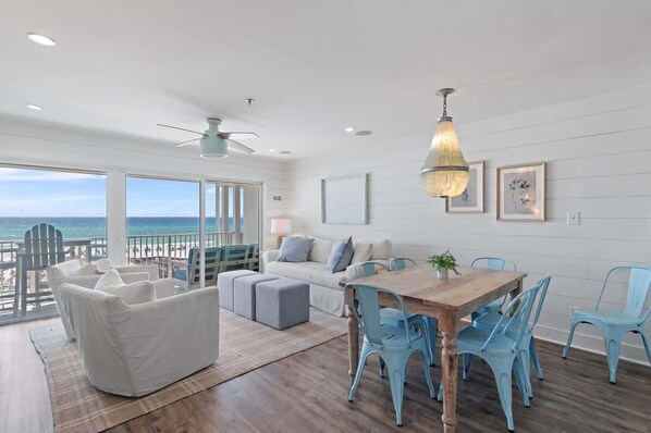 A spacious room with a large window overlooking the ocean, a ceiling fan, a dining table with chairs, and a cozy seating area.