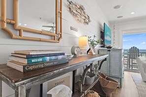 A room with a wooden table, books, a lamp, a framed picture, and a view of the ocean. - Seagrove - Commodore Retreat 202 (Santa Rosa Beach)