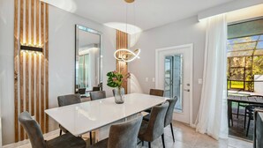 Een moderne eetkamer met een witte tafel, grijs gestoffeerde stoelen, een decoratieve vaas met bloemen en een grote spiegel.
