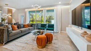 Een moderne woonkamer met een grijze bank, oranje poefjes, een salontafel en een grote televisie op een witte dressoir.