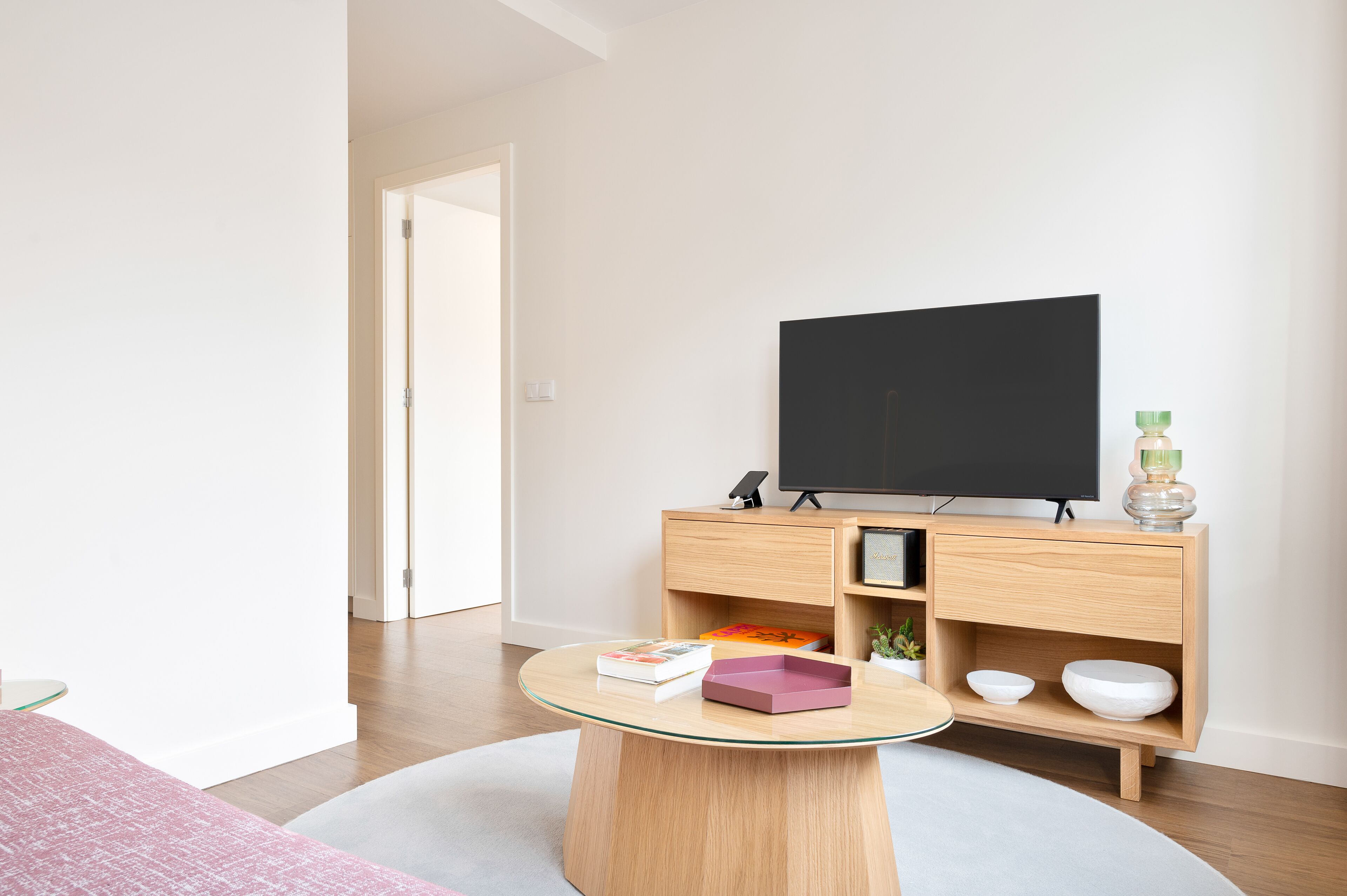 A modern living room with a round glass coffee table, a flat-screen TV on a wooden TV stand, and a minimalist decor.