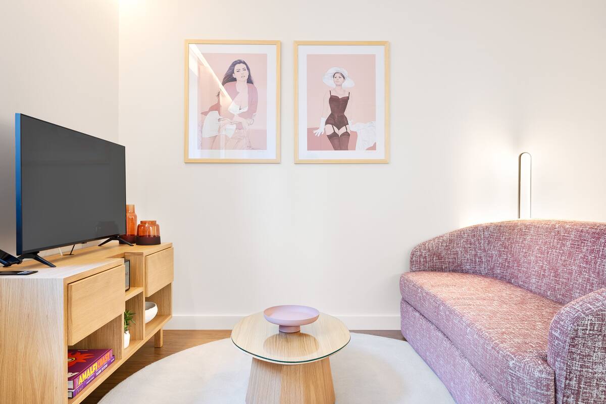 Een moderne woonkamer met een flatscreen televisie, een houten televisiemeubel, twee ingelijste kunstwerken, een ronde glazen tafel en een gestreept sofa.