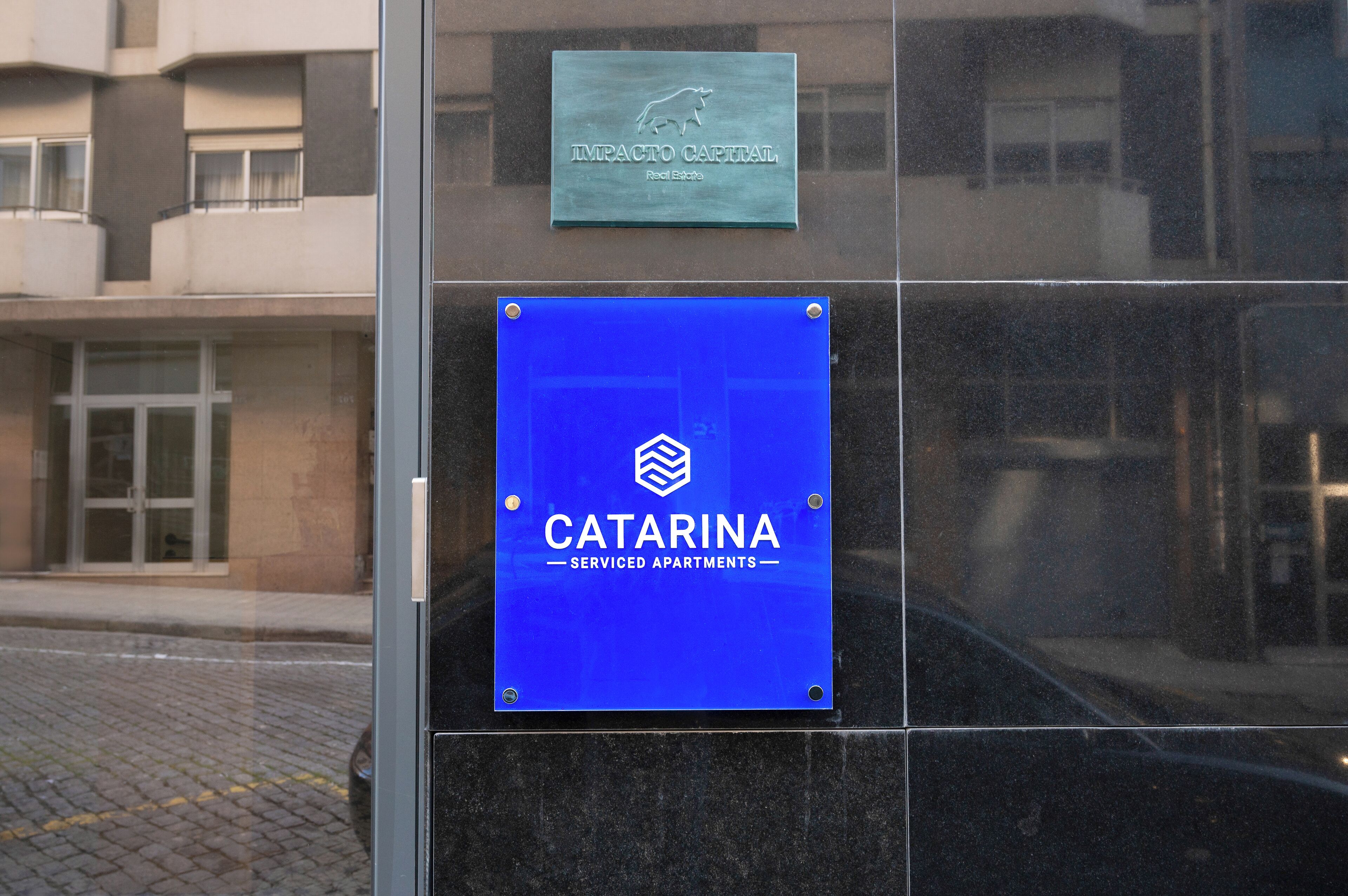 A building facade with two signs: one for 'IMPACTO CAPITAL Real Estate' and another for 'CATARINA Serviced Apartments'.