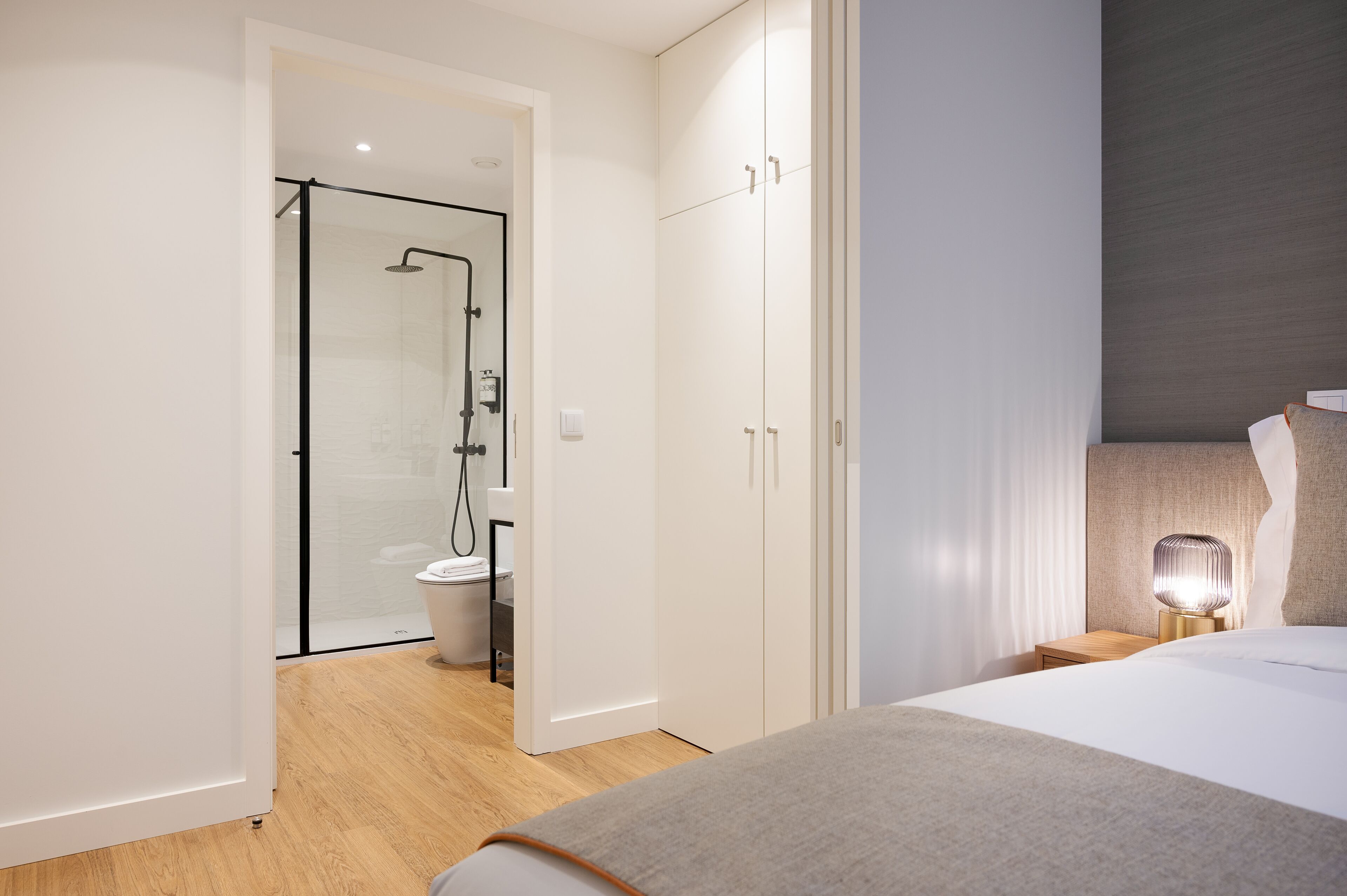 Ein modernes Hotelzimmer mit einem großen Bett, einem Nachttisch mit Lampe, einem Kleiderschrank und einer separaten Duschkabine.