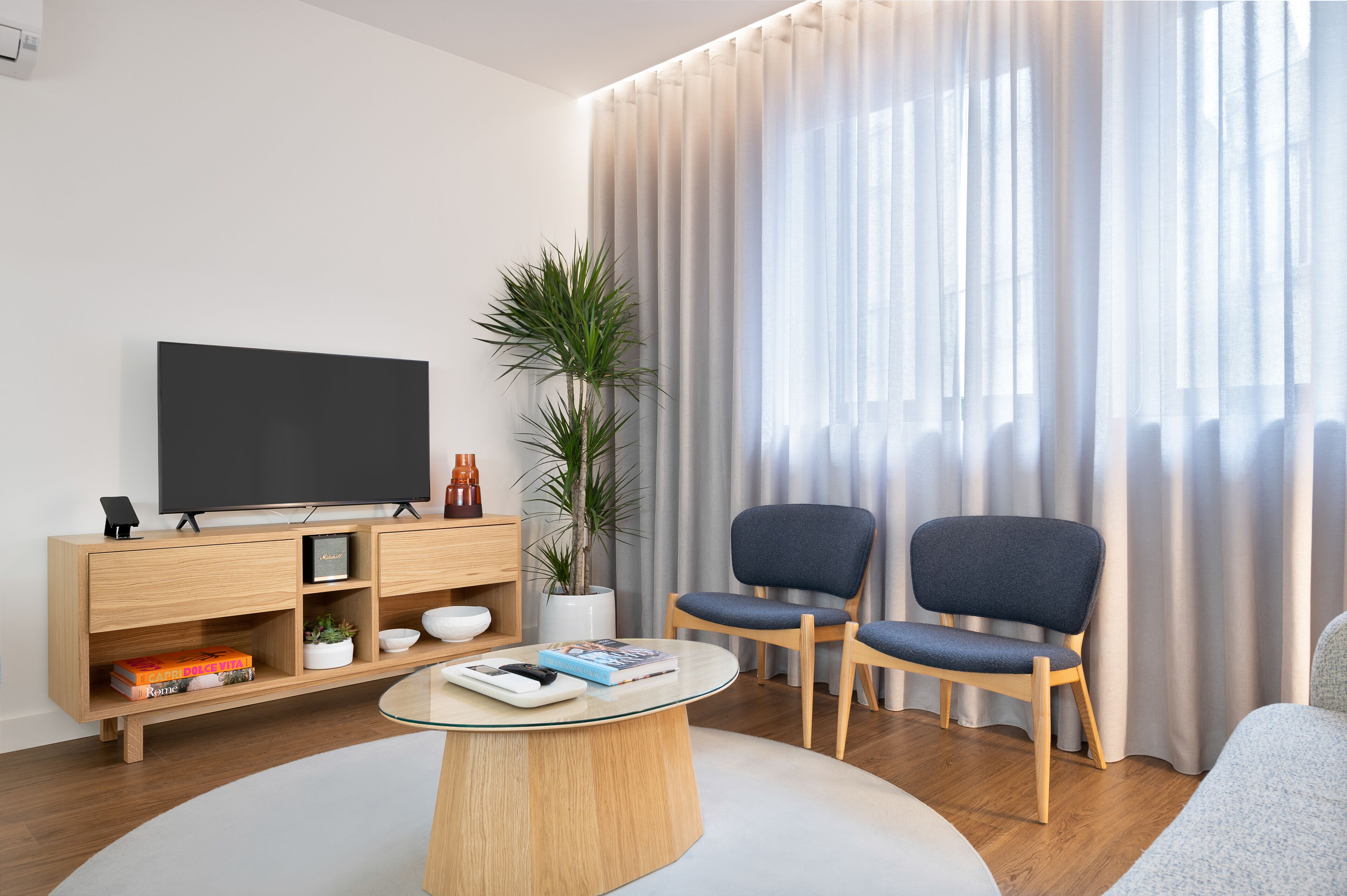 Ein modernes Wohnzimmer mit Flachbildfernseher, Holzmöbeln, einem Glastisch und einem großen Fenster mit transparenten Vorhängen.
