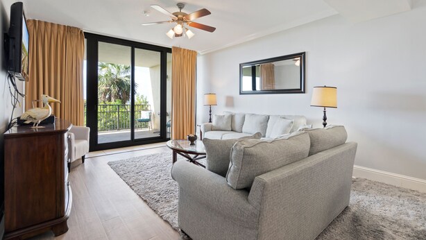 A living room with a grey sofa, a wooden cabinet, a ceiling fan, a TV, and a sliding glass door leading to a balcony. - Stay 2 nights and get an extra night free! - Unit 116 - Exceptions Apply (Dauphin Island)
