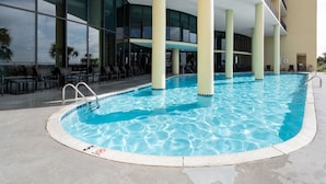 A swimming pool with a clear blue water surface, surrounded by a poolside area with chairs and tables, and a modern building with large windows in the background. - Stay 2 nights and get an extra night free! - Unit 116 - Exceptions Apply (Dauphin Island)