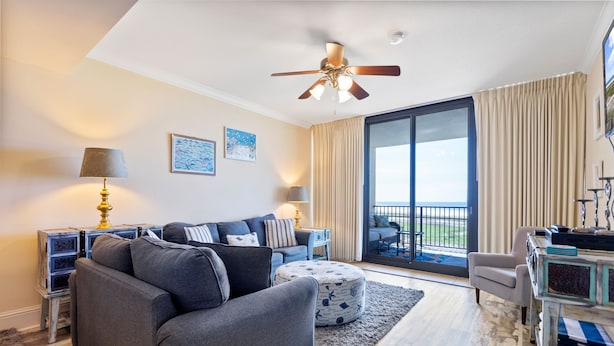 Un soggiorno moderno con un divano grigio, un poggiapiedi e una ventola a soffitto. Una grande finestra con tende, un tavolino con una lampada e vista sull'esterno.