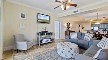 A living room with a grey sofa, a round ottoman, a white armchair, a wooden coffee table, a ceiling fan, and a TV mounted on the wall.