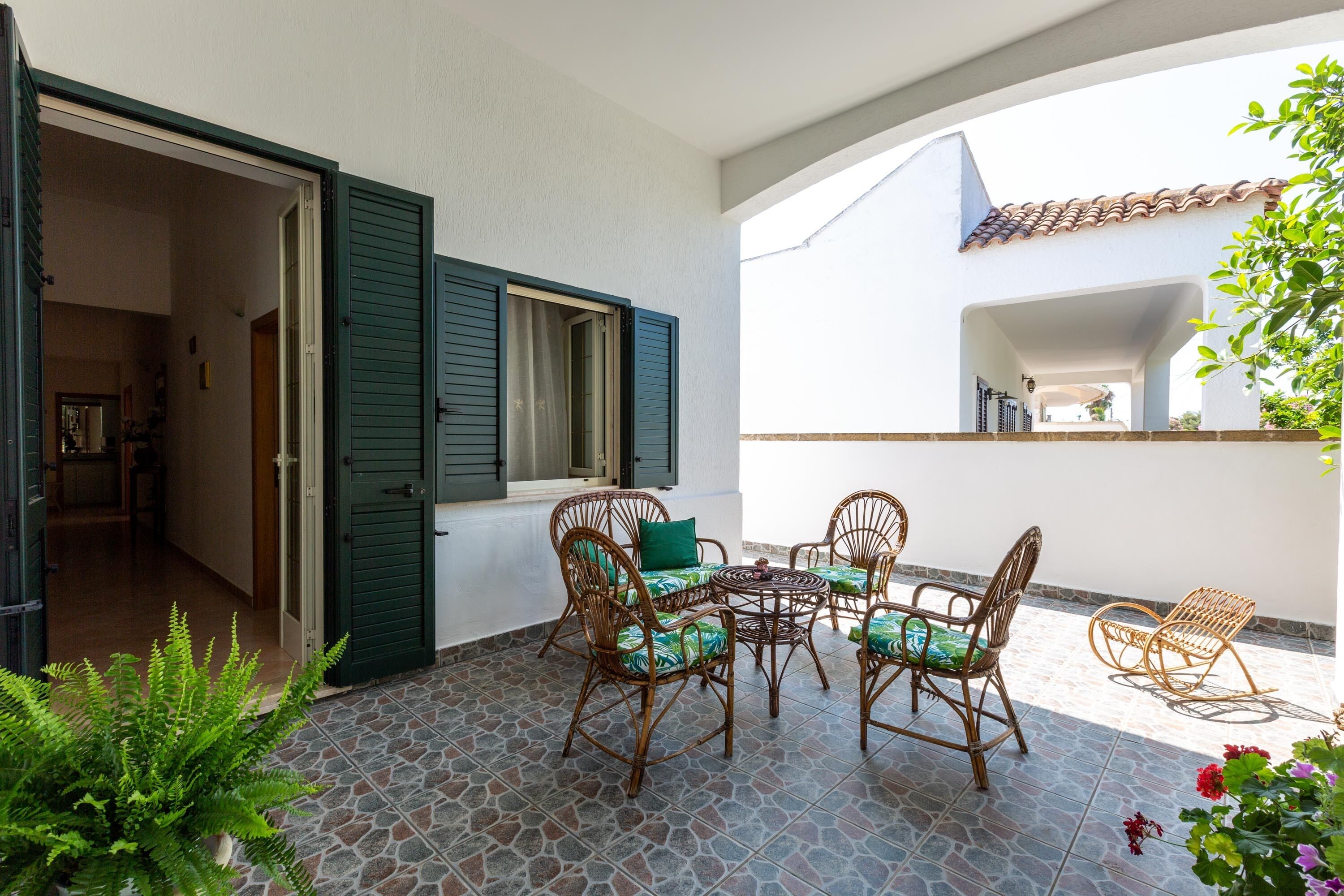An open patio with wicker furniture and a tiled floor, leading to a room with a door and window with shutters.
