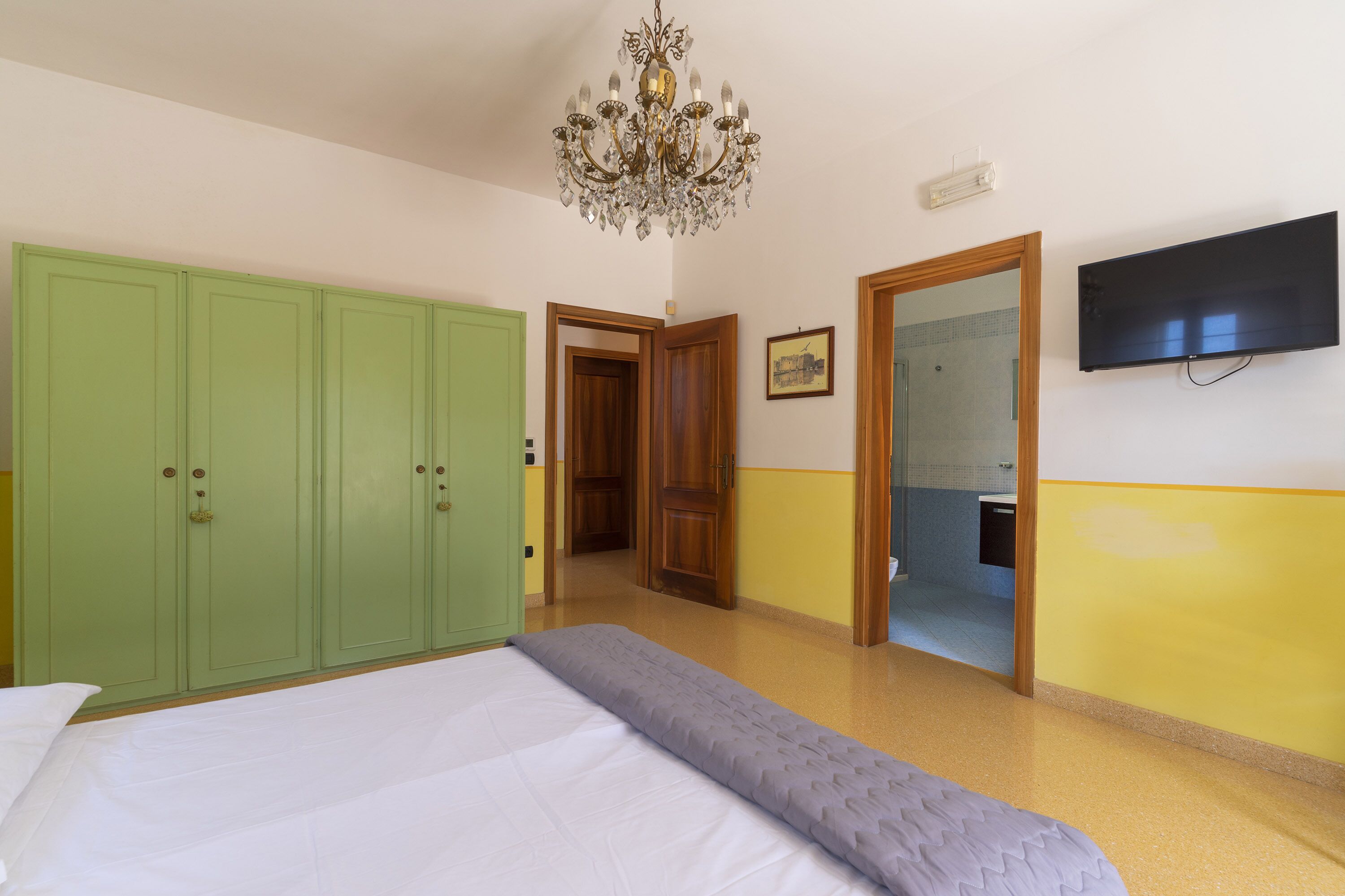 A bedroom with a bed, a green wardrobe, a television, and a chandelier.