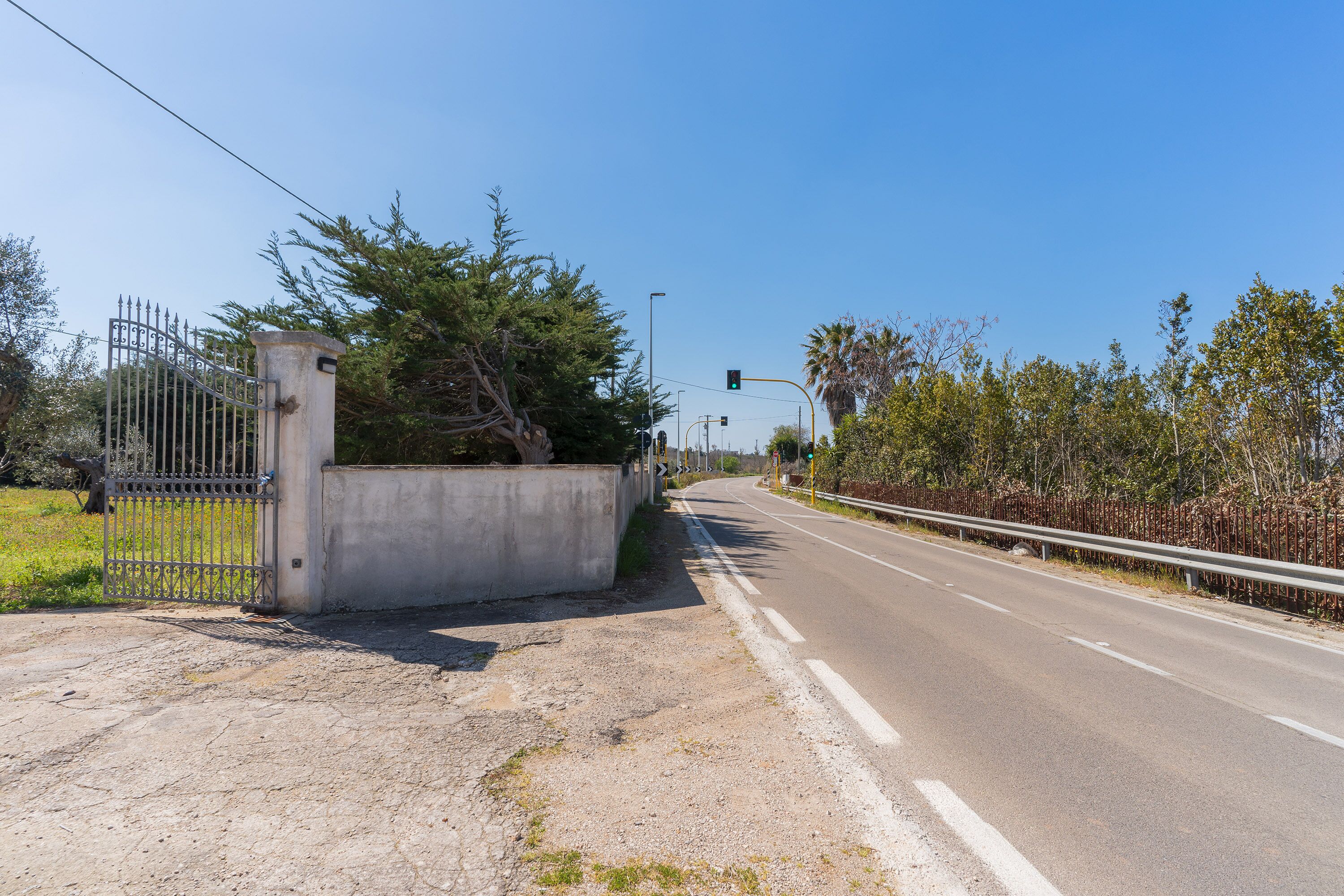A road with a metal gate and concrete wall on the left, surrounded by trees and a clear sky.