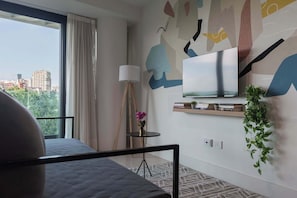 Ein modernes Hotelzimmer mit großem Fenster, einem Bett, einem an der Wand montierten Fernseher, einem kleinen Tisch mit Lampe und einem Regal mit Büchern und Pflanze.
