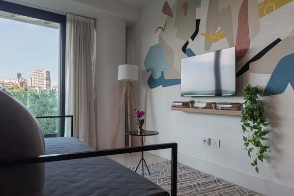 A modern hotel room with a large window, a bed, a TV mounted on the wall, a small table with a lamp, and a shelf with books and a plant.