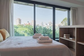 Ein Hotelzimmer mit einem großen Bett, einem Holfnachtkästchen und Blick auf die Stadt durch große Fenster.