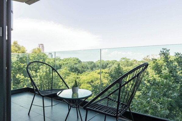 A balcony with two wicker chairs and a small table, overlooking a dense green forest and city buildings.