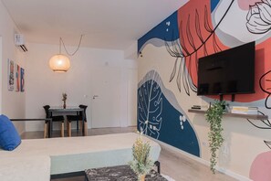 Un salon moderne comprenant un canapé, une table à manger avec des chaises, un luminaire suspendu, un téléviseur à écran plat fixé au mur et une fresque décorative.