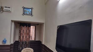 Ein Zimmer mit großem Fernseher, einem Fenster mit gemusterten Vorhängen, einem kleinen Tisch mit Stuhl und einem gerahmten Bild an der Wand.