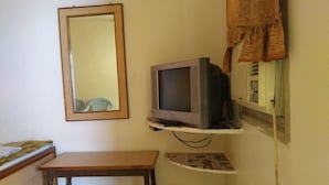 Ein kleines Zimmer mit einem Holztisch, einem Fernseher auf einem Regal, einem Spiegel an der Wand und einem Fenster mit Vorhang.