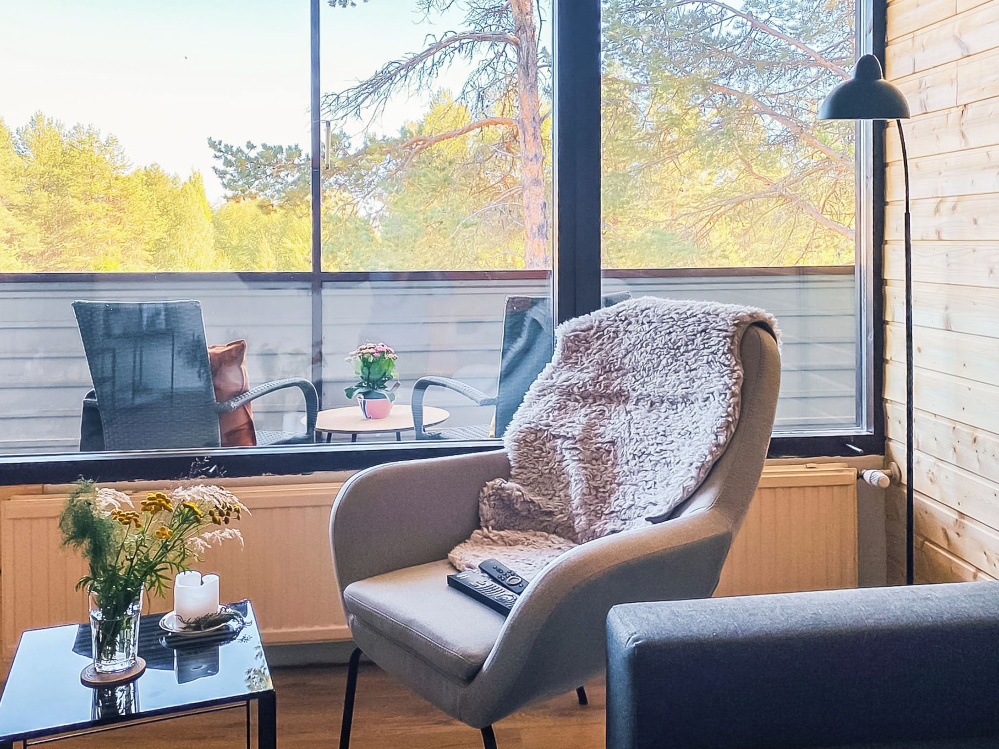 Una habitación acogedora con un amplio ventanal, un sillón cómodo, una mesita con un florero y vistas a árboles.