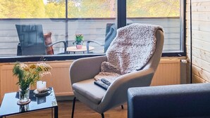 Una habitació acollidora amb un gran finestral, un butacó confortable, una taula petita amb un ram de flors i una làmpada.