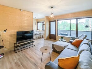 A modern living room with a flat-screen TV, wooden flooring, a sofa with cushions, a coffee table, and large windows offering a view of trees.