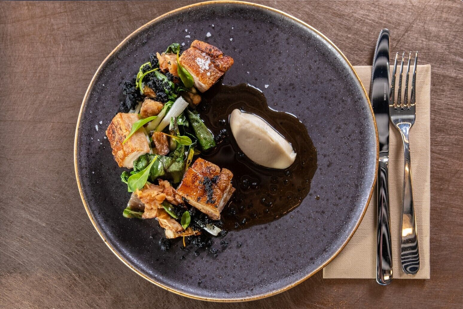 Un plato de comida con un plato de cerámica oscura, un tenedor y un cuchillo sobre una mesa de madera.