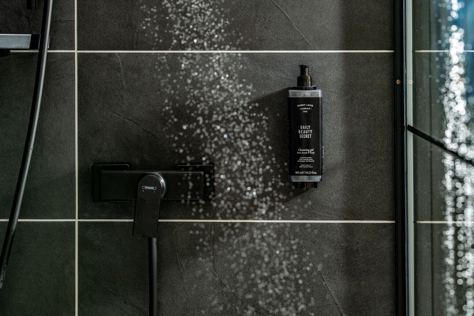 A shower area with a handheld showerhead and a bottle of Guest Love Daily Beauty Secret cleansing gel.