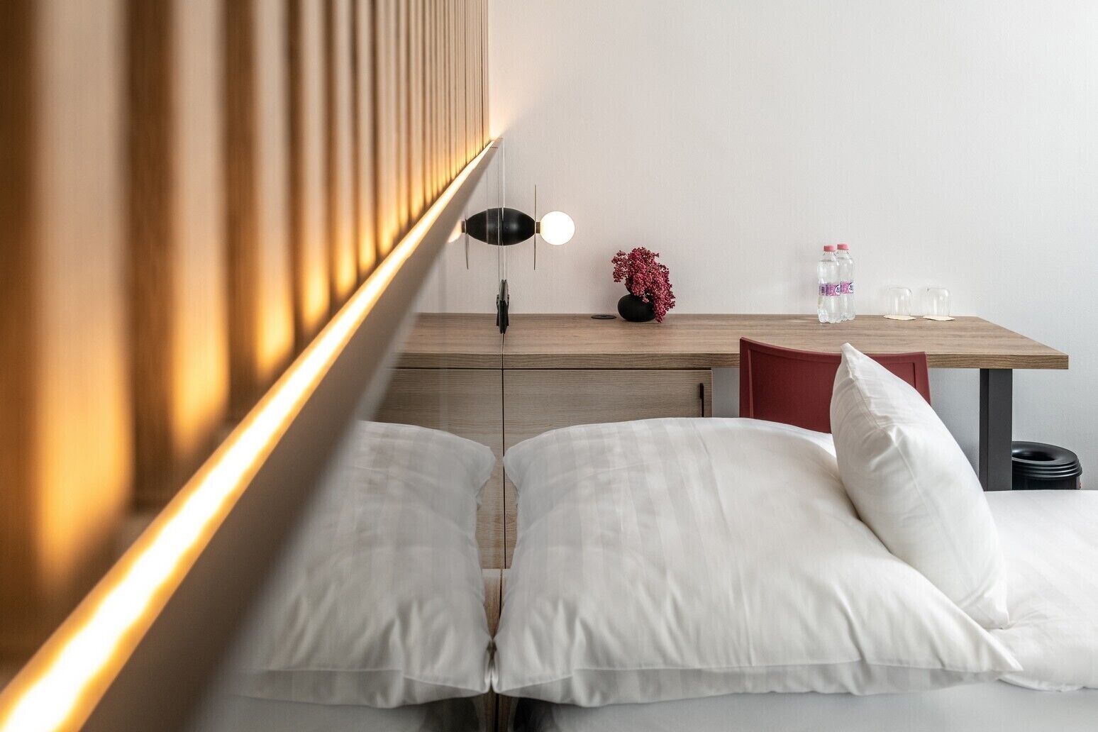 A modern bedroom with a bed, bedside table, lamp, and a small desk with a vase of flowers.