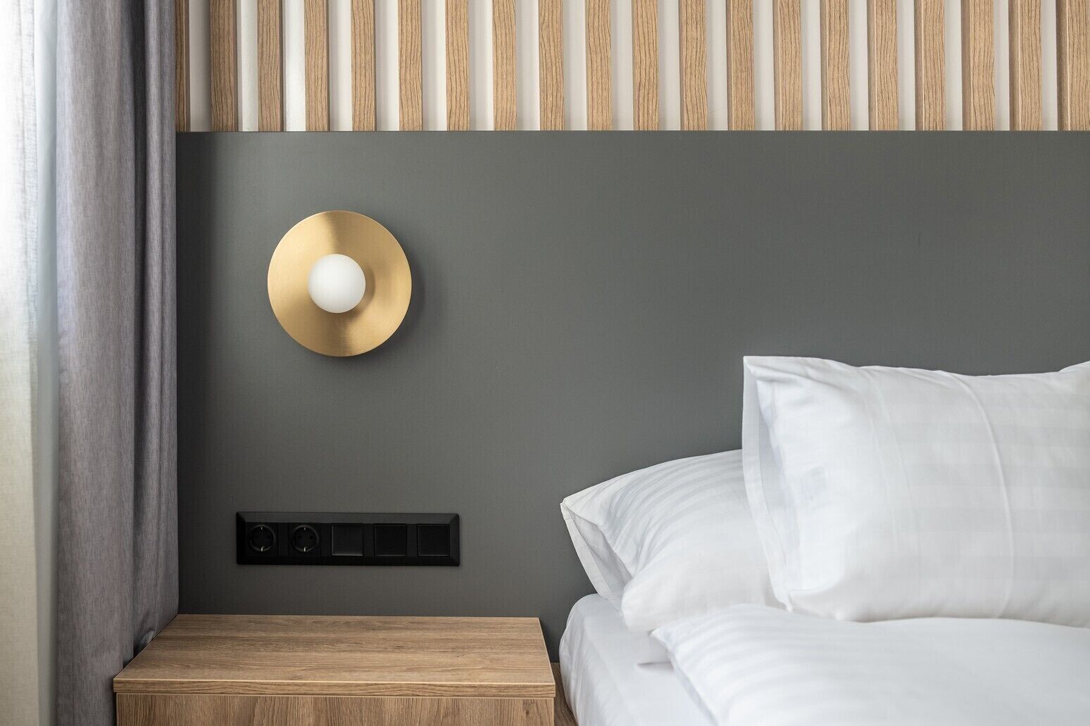 A modern bedroom with a grey wall, a wooden nightstand, a bed with white bedding, a round wall-mounted light, and a striped ceiling.