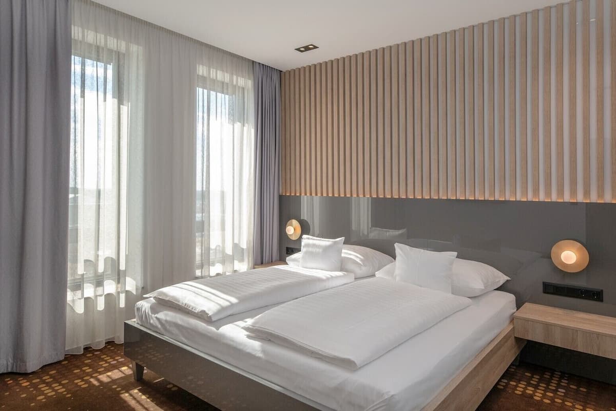 Una habitación de hotel moderna con una cama grande, dos mesitas de noche y vistas al exterior a través de cortinas translúcidas.