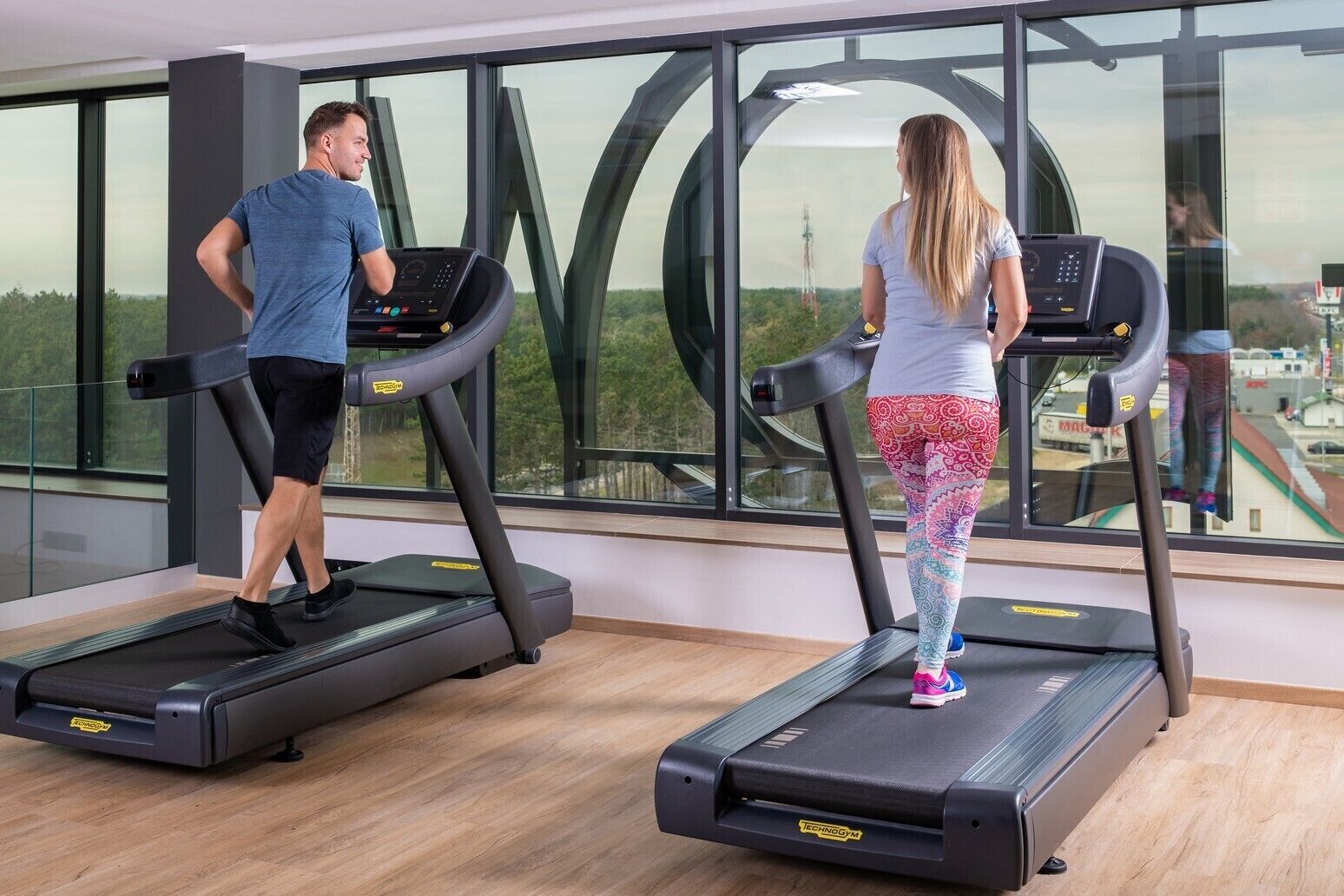Two people using treadmills in a gym with large windows and a view of a landscape.