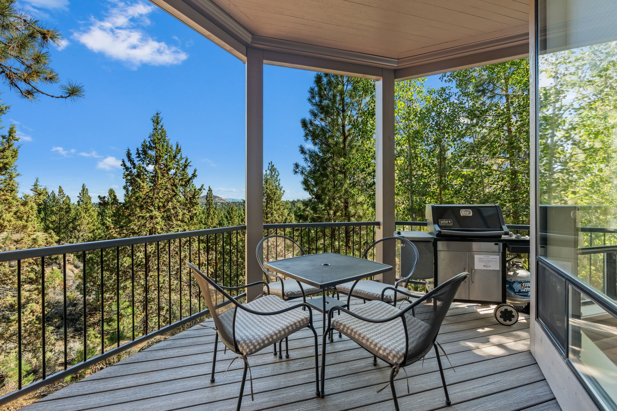 A covered outdoor area with a table and chairs, a grill, and a view of a forest.