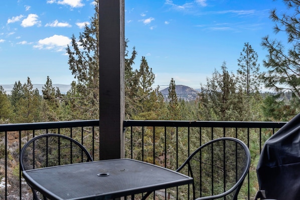 A balcony with a table and chairs, overlooking a forested area and a distant mountain range.