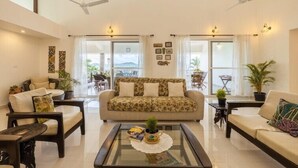 A spacious living room with a sofa, armchairs, and a coffee table. There are ceiling fans, large windows, and potted plants. - amã Stays & Trails Arcadia, Coorg (Virajpet)