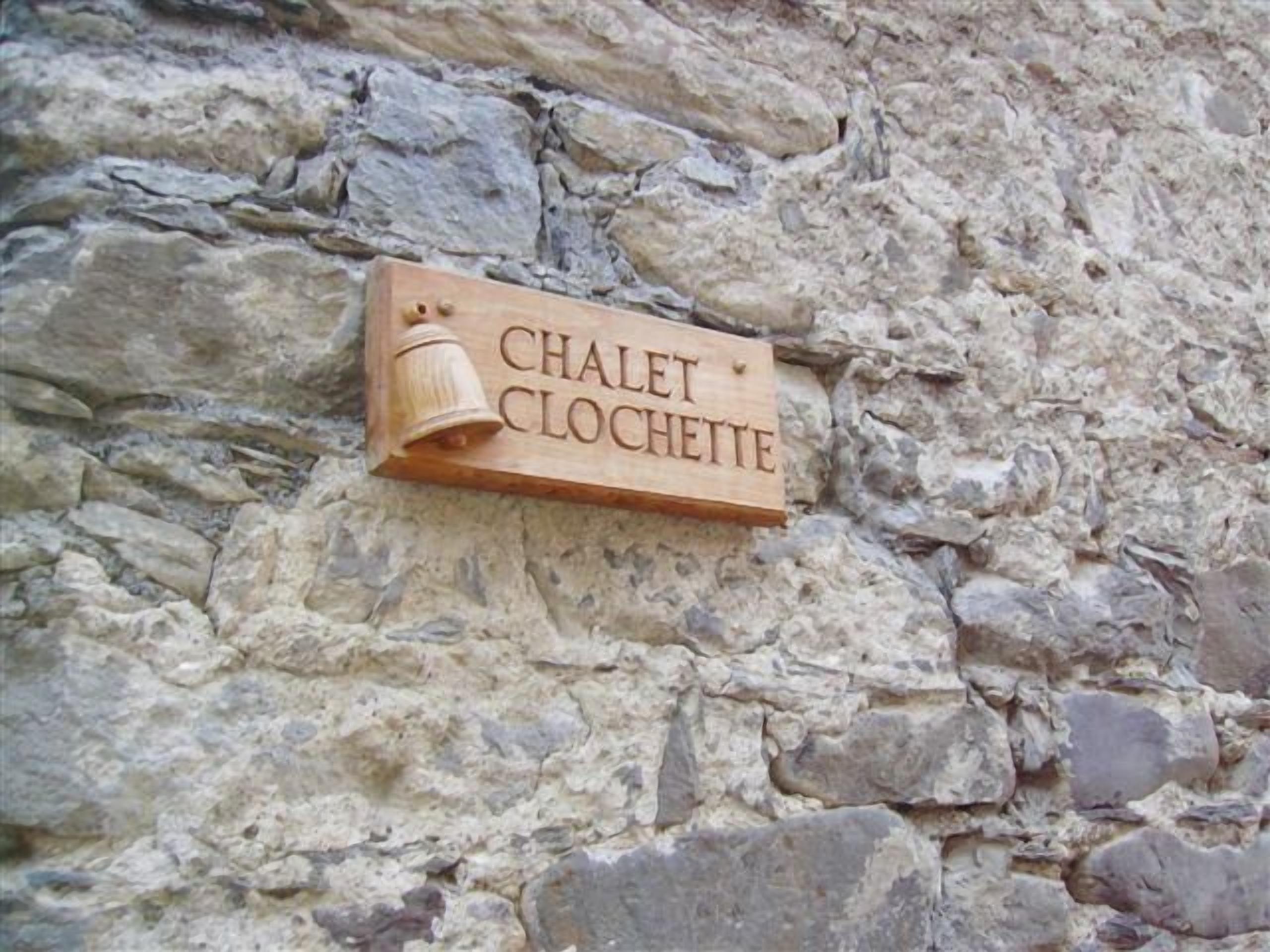 Une enseigne en bois portant l’inscription « CHALET CLOCHETTE », fixée à un mur de pierre.