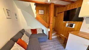 A cozy living area with a gray sectional sofa, wooden accents, a small kitchen area with a white refrigerator, and a staircase leading to an upper level. - Terrace, fireplace or stove, tv, ski locker, 30m², Les Menuires (Saint-Martin-de-Belleville)