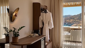 A hotel room with a view of a mountainous landscape, a wooden desk with a potted plant, a robe hanging on a rack, and a table with a coffee machine. - Armony Suites Milos (Milos)