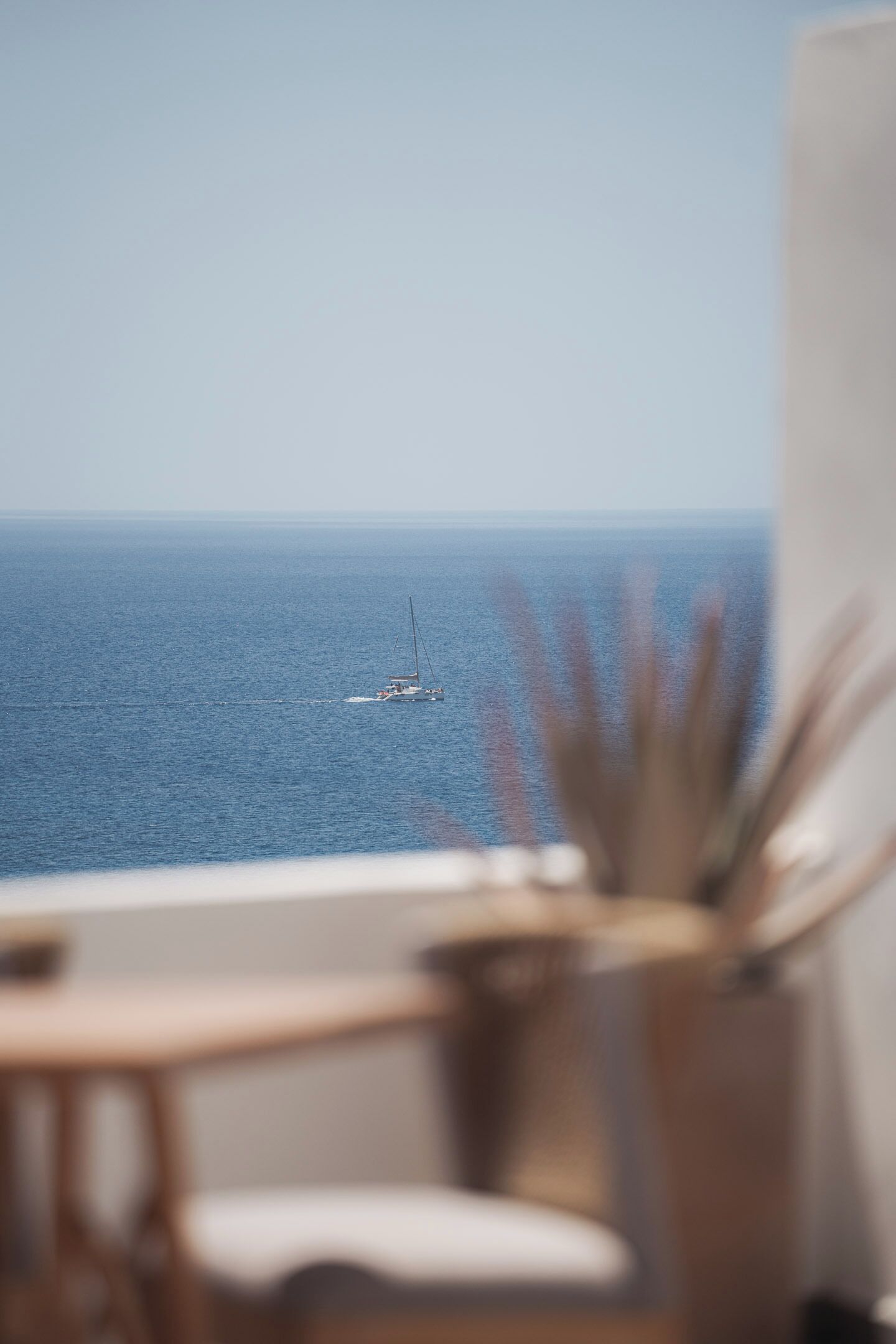 A view of the sea with a sailboat from a balcony with a chair and a plant.